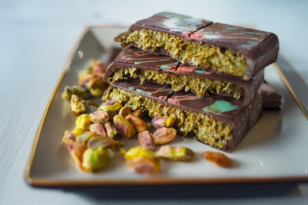 A plate of artisanal chocolate bars with a saffron and pistachio filling, accompanied by scattered pistachio nuts.How to get famous Dubai chocolate