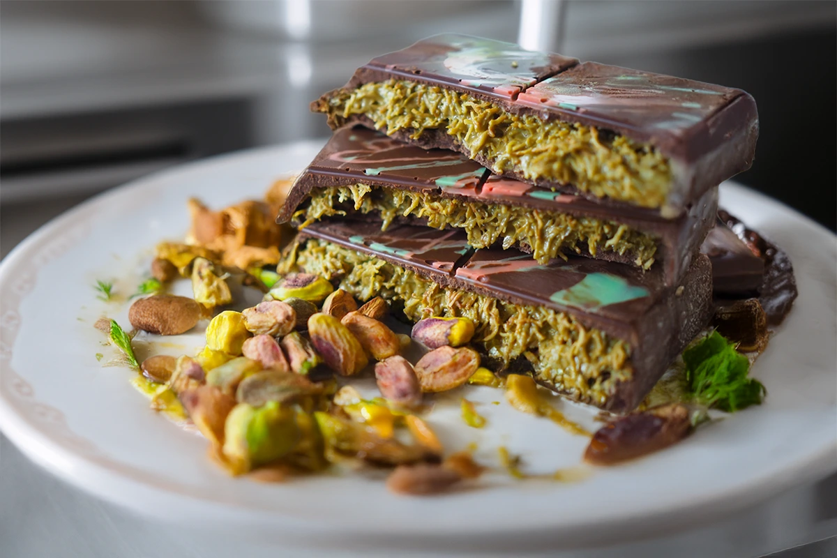 A stack of dubai chocolate bars recipe filled with pistachio-flavored filling, garnished with whole pistachios and herbs on a white plate.