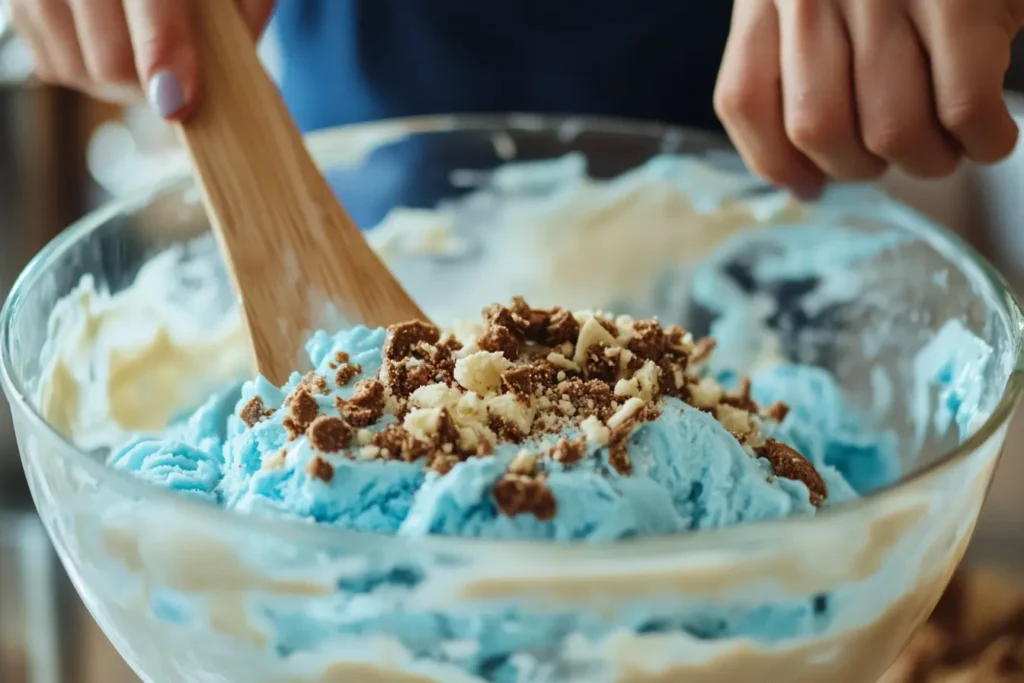 A person mixing bright blue Cookie Monster Ice Cream in a glass bowl with a wooden spoon, topped with crushed chocolate and vanilla cookies.